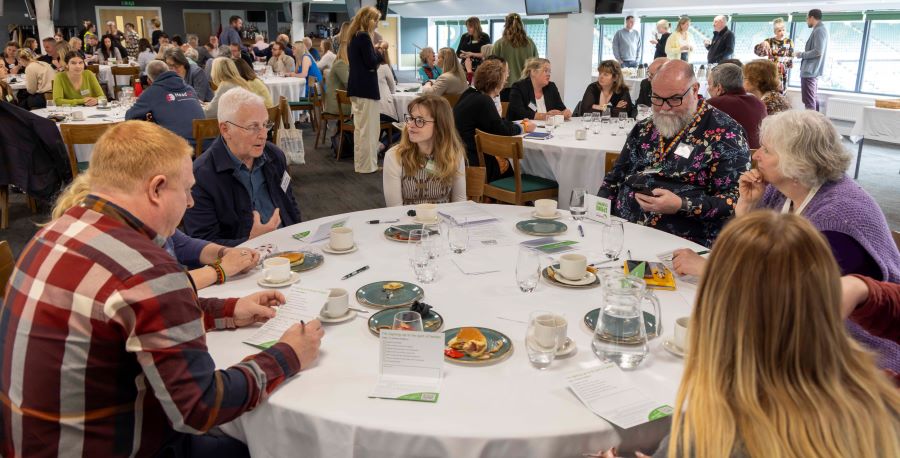 A group of people sit around a table at an event, enagged in conversation over coffee and breakfast. Behind is a busy event, with a view of Home Park stadium beyond.
