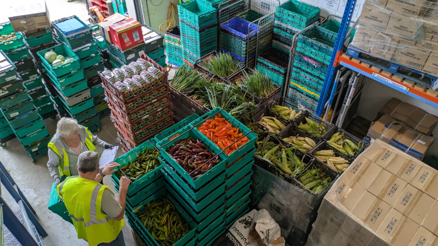 A warehouse full of stacked food pallets and crates, with two volunteers in hi vis.
