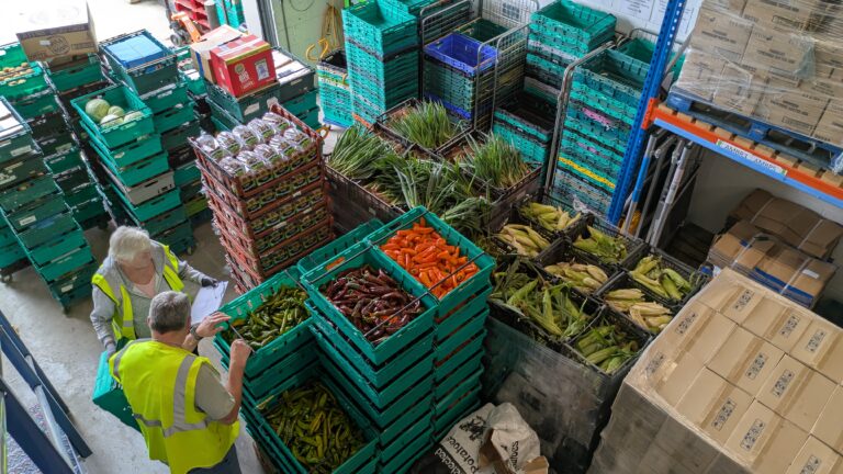 A warehouse full of stacked food pallets and crates, with two volunteers in hi vis.