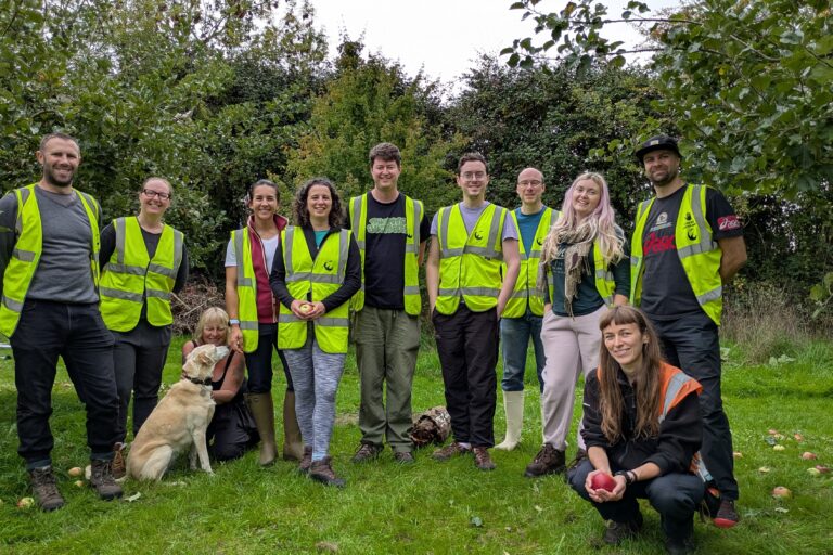 Group of people wearing high vis vests stand or crouch in a group. All are looking at the camera and smiling. A pale brown dog is also sat with them. They are in an orchard with trees in the background