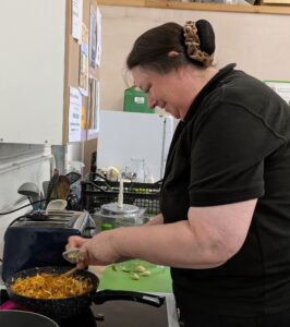 Woman wearing a black polo shirt is grating garlic into a pan full of veg on a hob. There are cupboards and a fridge in the background