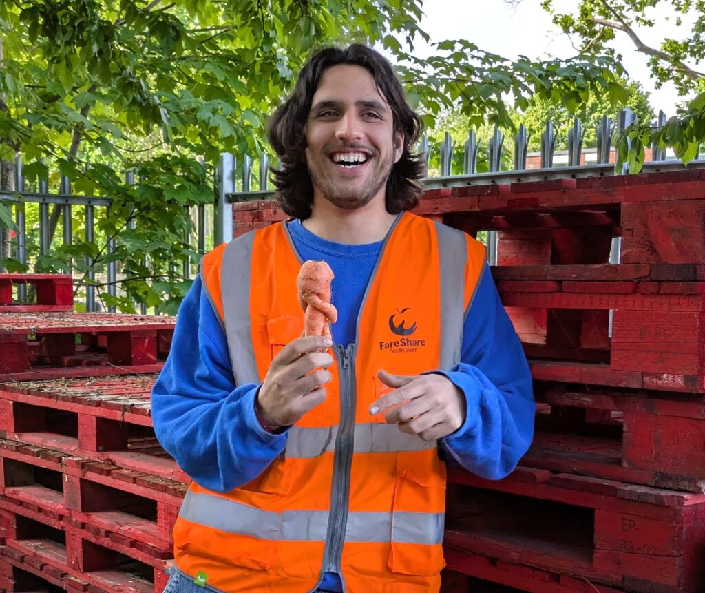 Robin is outside surrounded by trees and empty wooden pallets. He's got long dark hair and is wearing a high vis and laughing as he holds up a carrot.