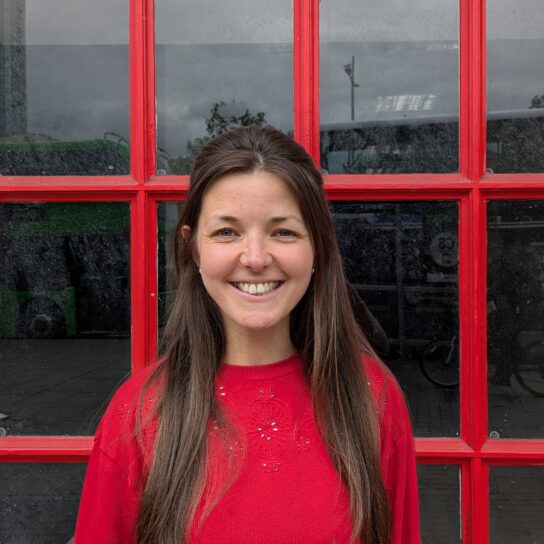 Woman with dark hair and a red top stands in front of a large window with red window panes. She is smiling straight at the camera