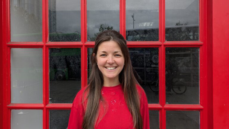 Woman with dark hair and a red top stands in front of a large window with red window panes. She is smiling straight at the camera