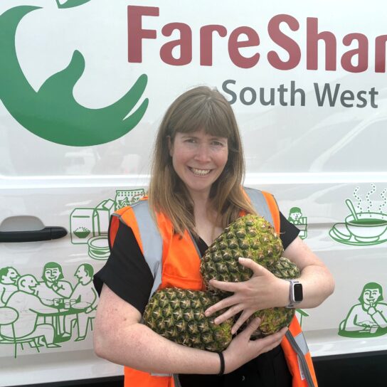 Woman with brown hair wearing an orange high vis stands in front of a FareShare South West branded van. She is holding three large pineapples and smiling
