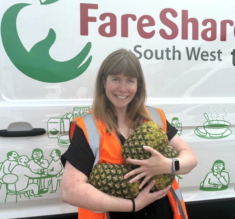 Woman with brown hair wearing an orange high vis stands in front of a FareShare South West branded van. She is holding three large pineapples and smiling