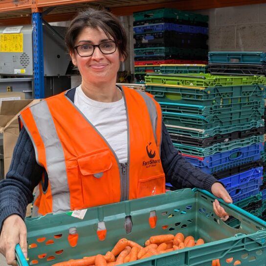 Woman with dark hair and glasses wearing an orange high vis vest stands holding a green crate full of carrots. She is standing in a warehouse and there are pallets stacked behind her