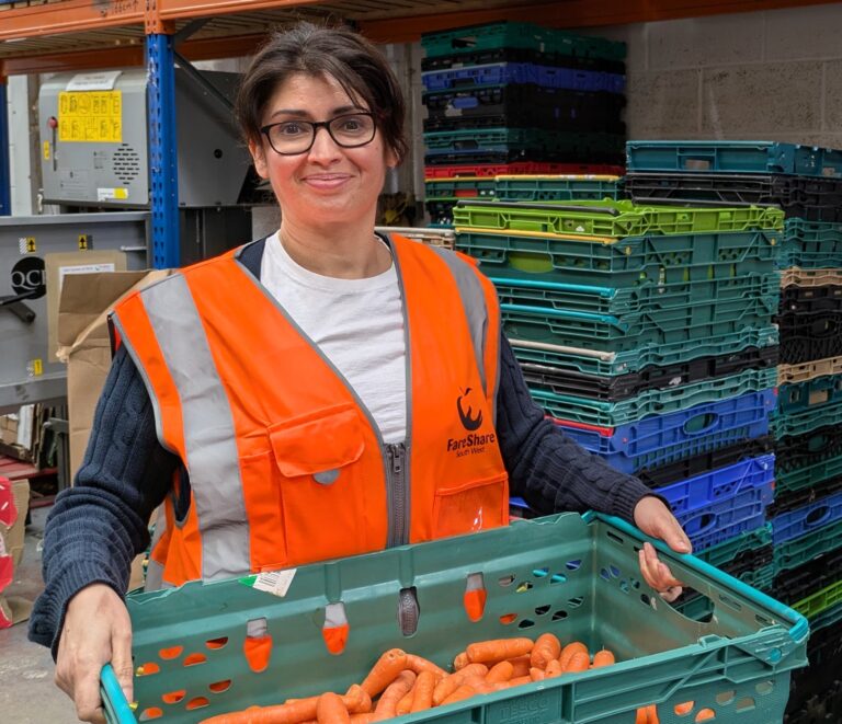 Woman with dark hair and glasses wearing an orange high vis vest stands holding a green crate full of carrots. She is standing in a warehouse and there are pallets stacked behind her