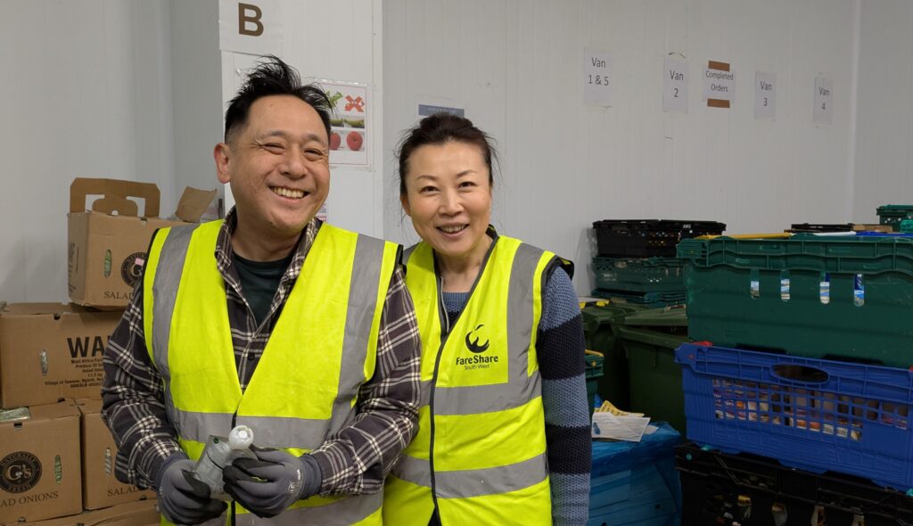 A man and a woman, both wearing yellow high vis vests, smile at the camera. They are in a chiller with stacks of crates surrounding them