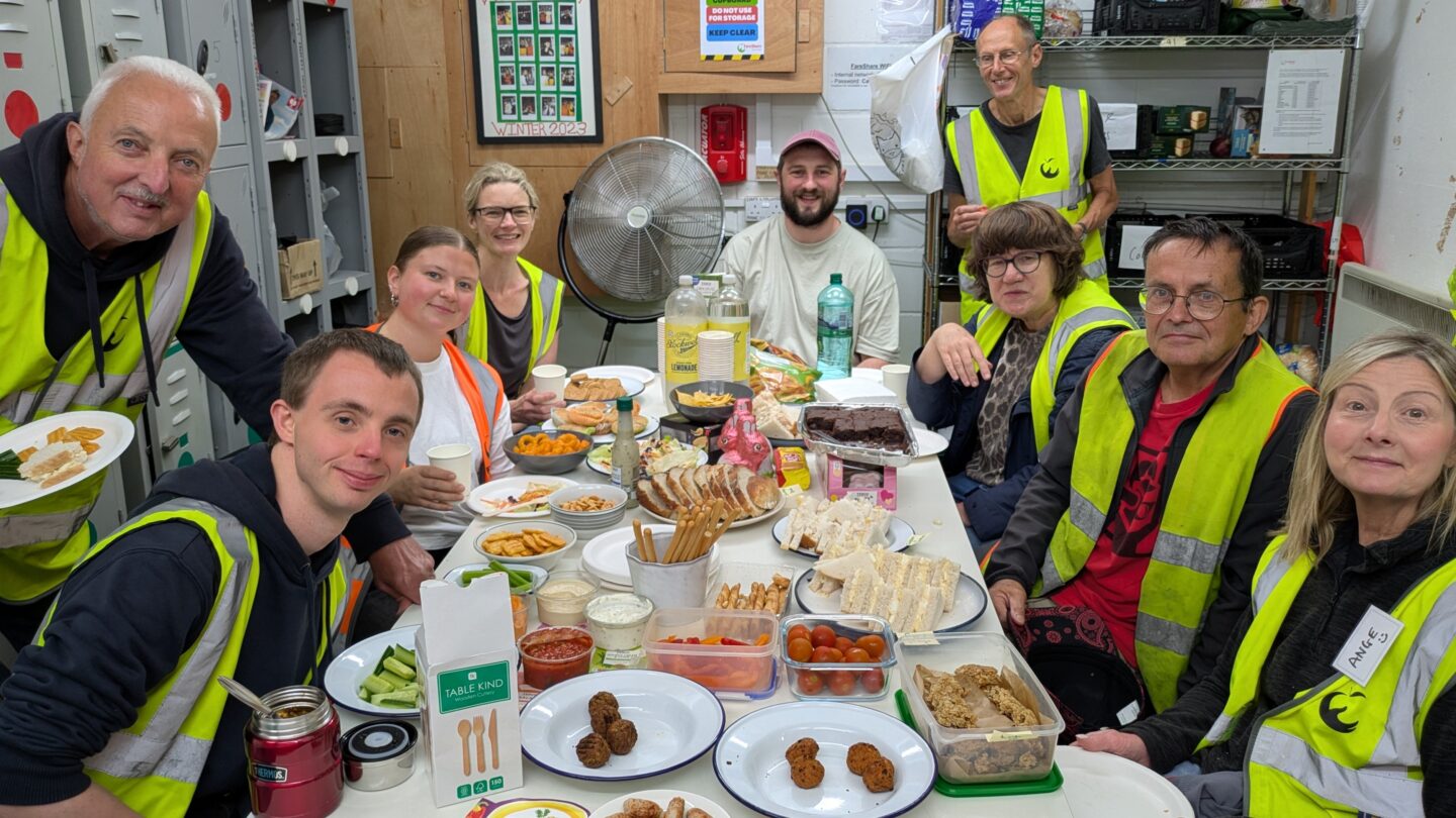 Group of people wearing yellow high vis vests are gathered around a table laden with food. They are in a work kitchen environment and are all looking at the camera and smiling