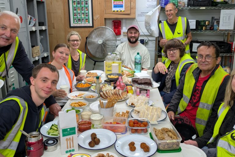 Group of people wearing yellow high vis vests are gathered around a table laden with food. They are in a work kitchen environment and are all looking at the camera and smiling