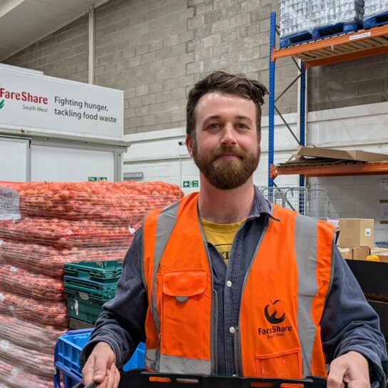 Man with dark hair is wearing an orange high vis vest. He is standing holding a crate in a warehouse, with pallets of food behind him