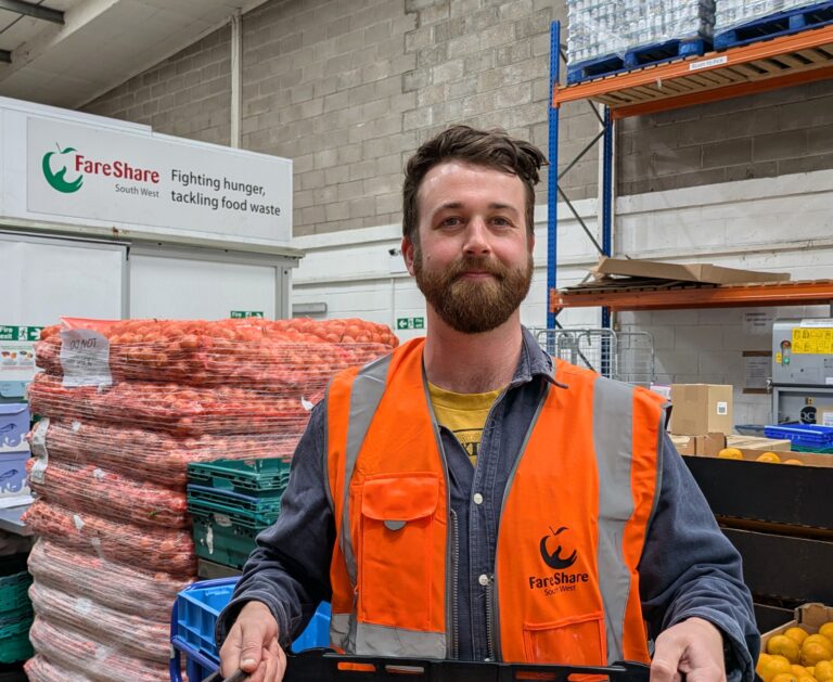 Man with dark hair is wearing an orange high vis vest. He is standing holding a crate in a warehouse, with pallets of food behind him
