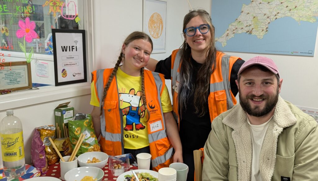 Two women wearing orange high vis vests stand behind a man wearing a cream jacket and pink cap. They are next to a table with leftovers of a party food spread on it. There is a window to one side and walls with a framed print and large map behind them. All are smiling at the camera