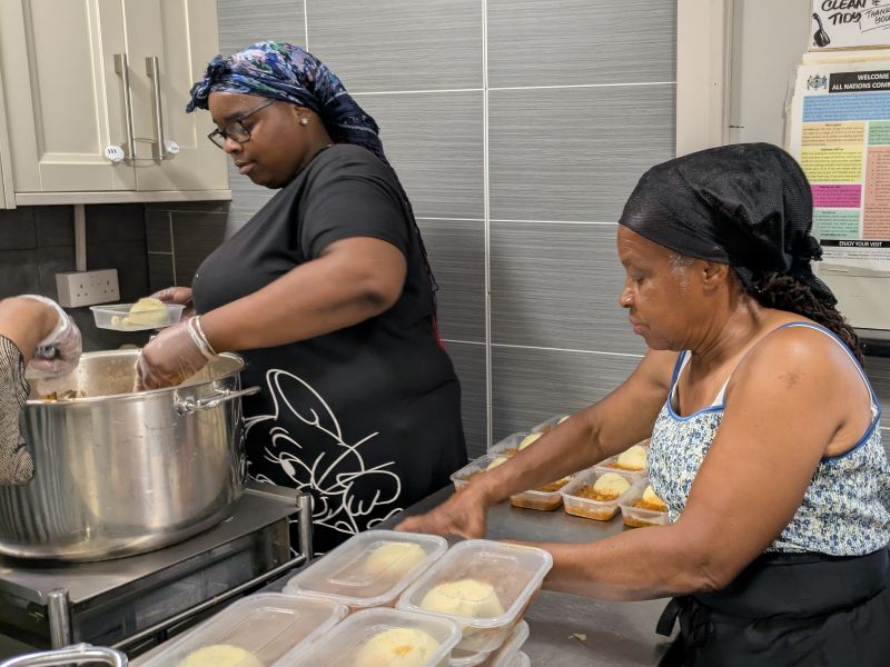 Rochel and Daphne in the kitchen: two women are cooking, one stirring a huge pan on the stove, one adding portions to containers.