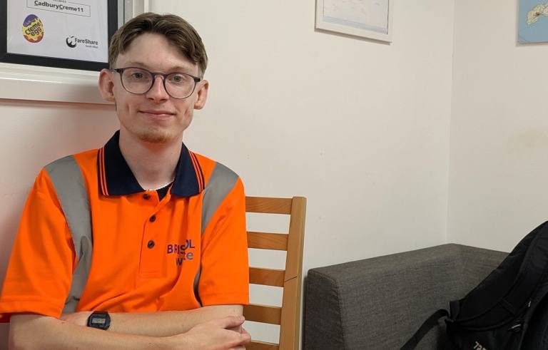 Young man wearing glasses and a bright orange, Bristol Waste branded uniform sits at a table as he smiles at the camera. There is a pale wall and the corner of a dark grey sofa behind him. There is also a framed print just behind his head