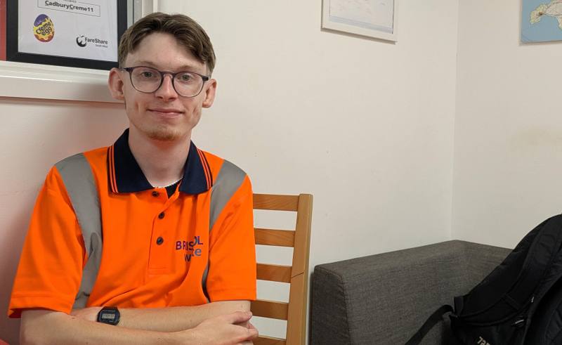 Young man wearing glasses and a bright orange, Bristol Waste branded uniform sits at a table as he smiles at the camera. There is a pale wall and the corner of a dark grey sofa behind him. There is also a framed print just behind his head