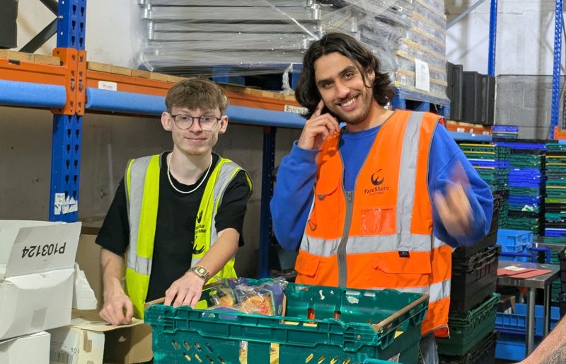 Two men smile at the camera as they stand side by side. The man on the left wears glasses and a black t-shirt with a yellow high vis vest over it. The man on the right wears a bright blue jumper with an orange high vis vest over it. They are in a warehouse and have a green crate and some packaged food in front of them