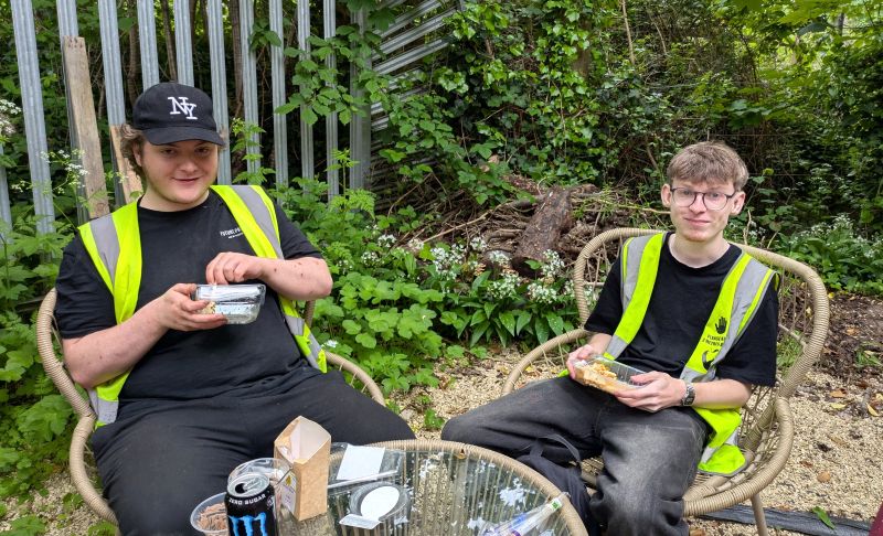 Two men sit on wicker-style chairs outside. Both are wearing black tops with yellow high vis vests over them. The man on the left has a black cap on. Both are smiling at the camera and holding a plastic tray of food. There is a table in front of them with some empty food and drink packaging. There are trees, foliage and a metal fence behind them