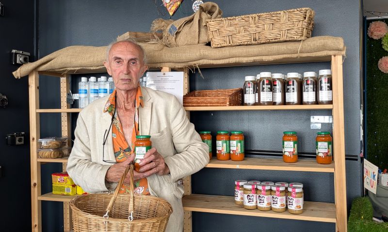 Andrew is an older white man, smartly dressed. He's holding a wicker shopping basket and a jar of pasta sauce in a community shop. Behind him is a dark grey wall and wooden shelves full of food.