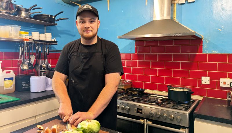 Morgan is wearing a baseball cap and a black apron in a brightly coloured kitchen. He's chopping fresh vegetables and looking directly at the camera with a soft smile.