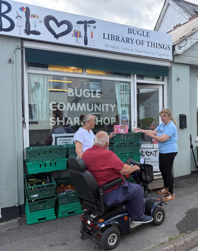 Photo of three people outside a shop. One, standing up, is wearing a blue t-shirt and black leggings - she is gesturing towards some crates. Another person is standing near her wearing a white shirt. They are talking to someone on an electric mobility scooter wearing a red shirt and blue trousers. 

Writing on the shop window says Bugle Library of Things. There are crates of fresh vegetables on display outside the shop