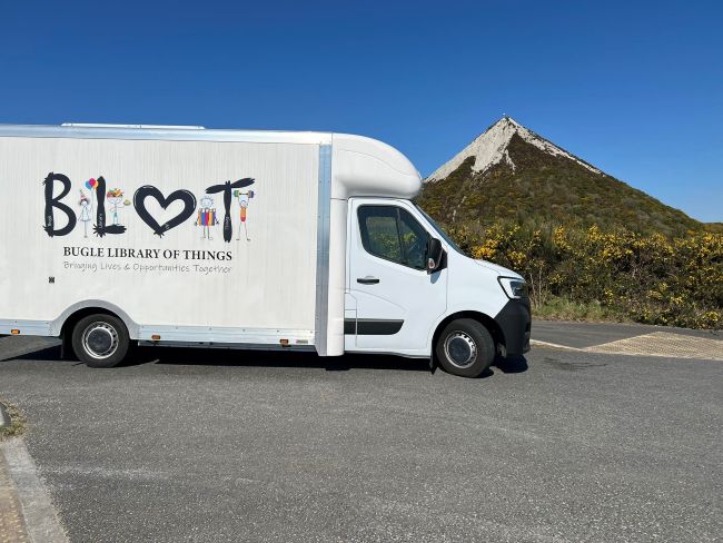 Photo of a white van with a large Bugle Library of Things logo on the side. The image is superimposed in front of a mountain background with blue sky