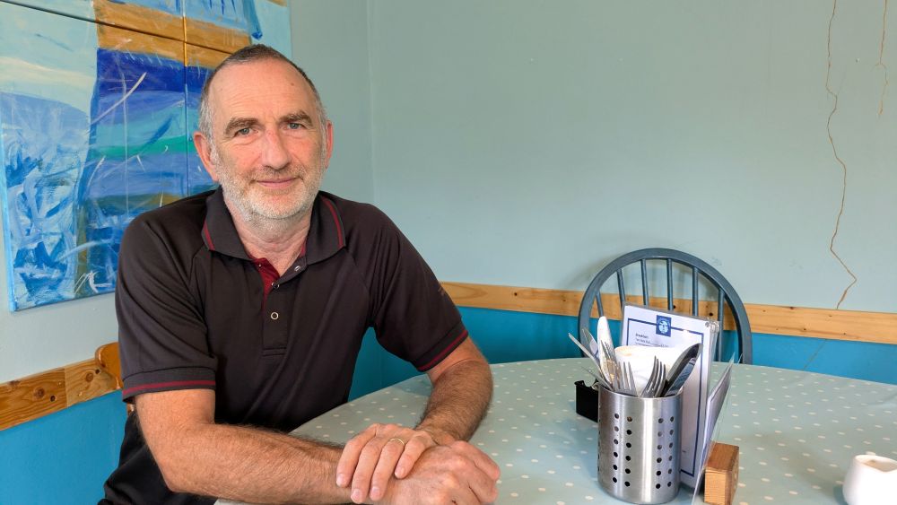 Man wearing a black polo shirt smiles at the camera as he sits at a table. There are blue walls with a large picture behind him and the table has a blue and white spotty table cloth, with a pot of cutlery and a menu on top