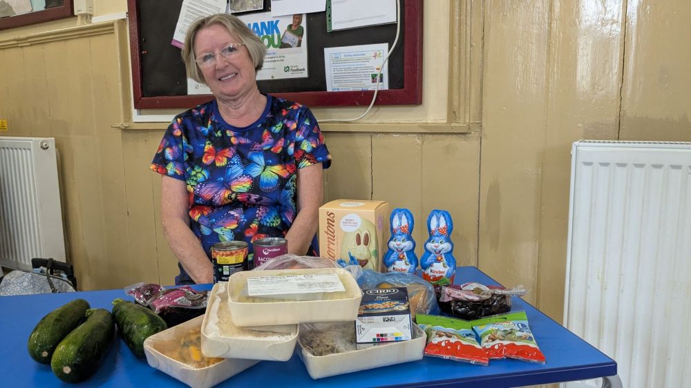 A woman with short hair and glasses, wearing a bright t-shirt with a distinctive butterfly design sits behind a table. On top of the table is a collection of food including chocolate bunnies, courgettes, wrapped beetroot, and a carton of chickpeas.