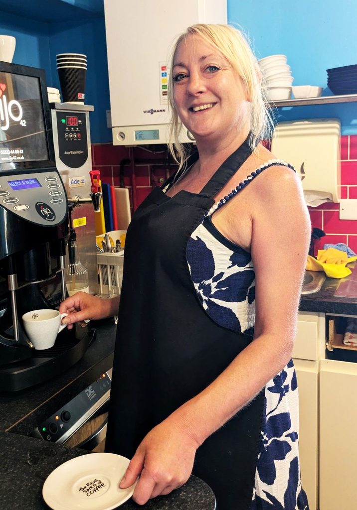Woman wearing a blue and white flowery summer dress with a black apron on top smiles at the camera as she holds a white cup under a coffee machine. She is standing in a kitchen and there are kitchen utensils and a red tiled wall behind her