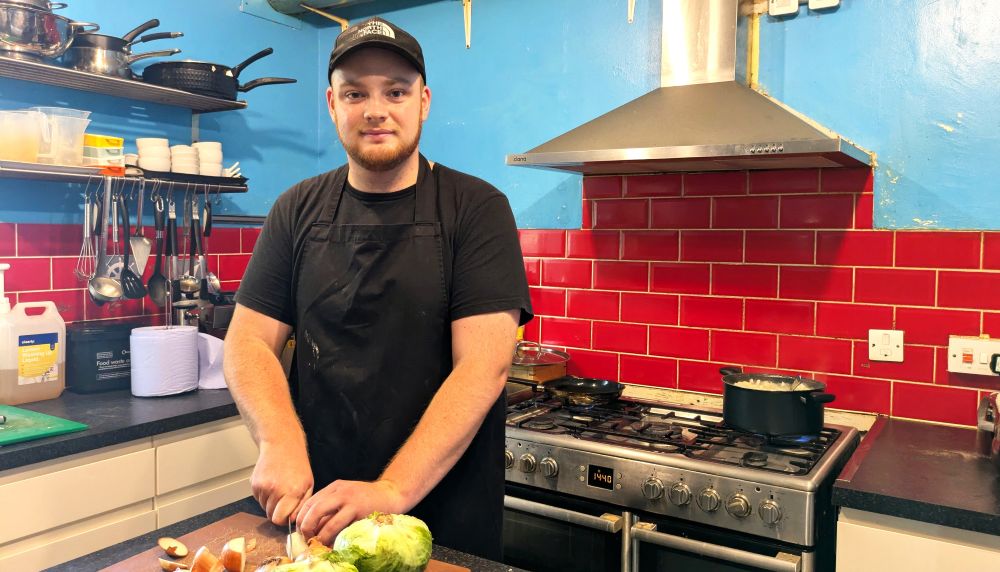 A young man wearing a black t-shirt, black cap and black apron looks at the camera. He is mid-way through chopping vegetables. He is standing in a kitchen and there are kitchen utensils, a hob and blue walls with red tiles behind him