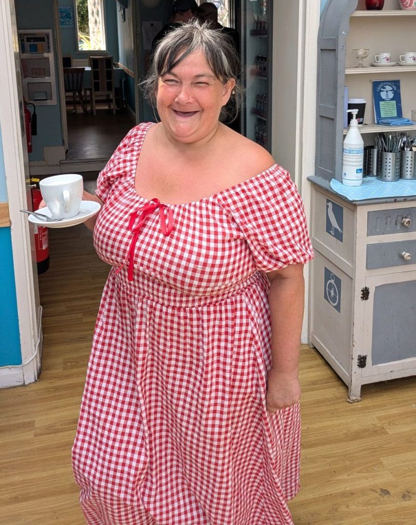 Woman wearing a red and white checked dress is holding a white cup and saucer and smiling at the camera. She is standing in a cafe setting and there is a dresser with pots of cutlery behind her