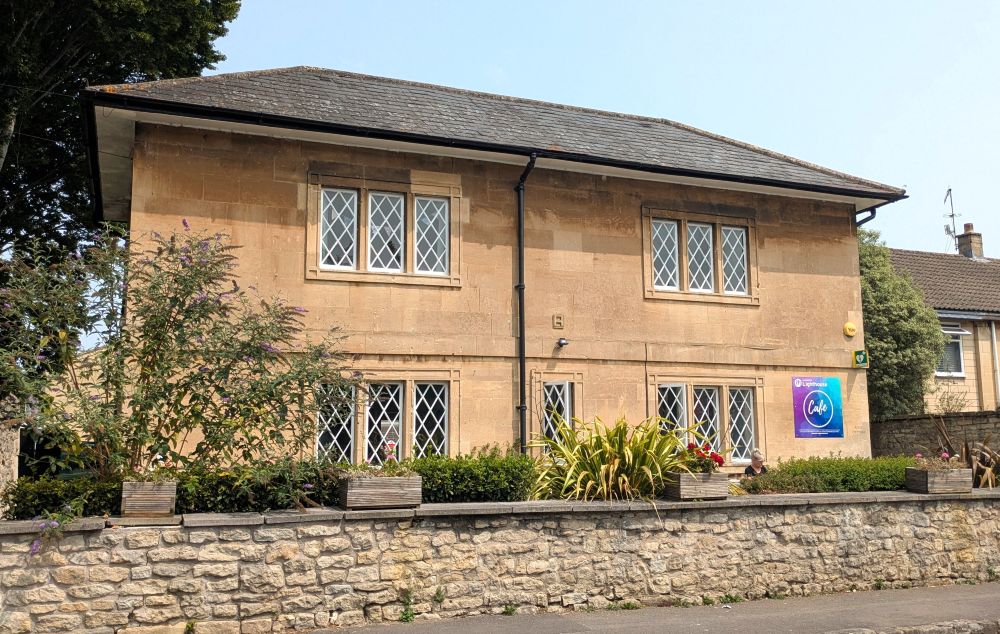 An attractive stone building with ornate windows. It has a wall out the front, with foliage visible behind the wall, a tree to the left hand side and blue sky above. There is a purple and blue sign on the side of the building that says 'cafe' in big font and lighthouse written smaller