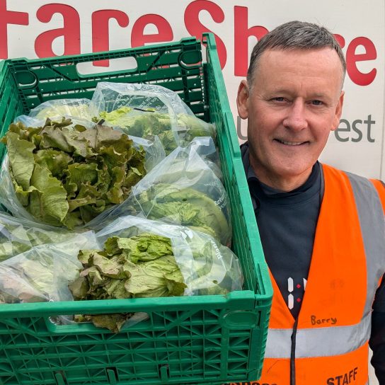 A man wearing a dark top underneath an orange high vis vest smiles at the camera as he holds a crate of green lettuces over one shoulder. He is standing in front of a FareShare South West-branded van