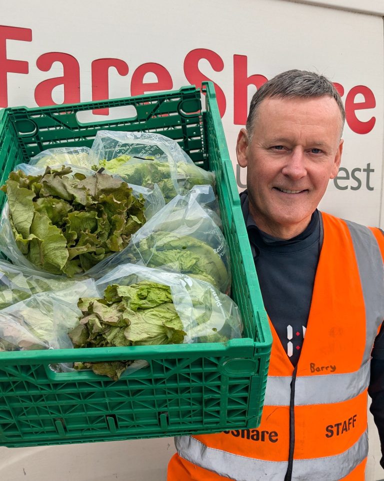 A man wearing a dark top underneath an orange high vis vest smiles at the camera as he holds a crate of green lettuces over one shoulder. He is standing in front of a FareShare South West-branded van