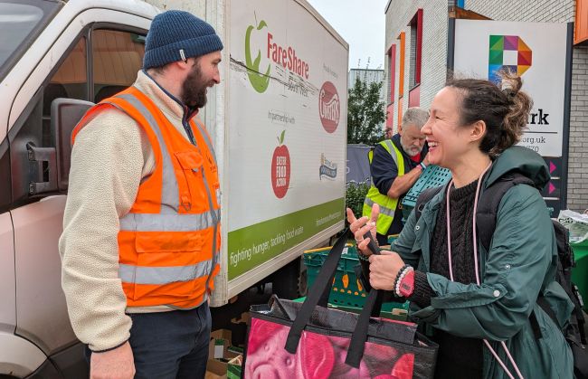 Woman wearing a black top underneath a dark green coat smiles as she holds a large shopping bag. She is looking towards a man wearing a beige fleece underneath an orange high vis vest and a blue woolly hat. There is a van with a FareShare South West logo, among other business logos on the side, behind him and there is another man wearing a yellow high vis vest lifting a green crate in the background. In the background to the right of the photo is the edge of a building and a large sign on which   the end of the word 'The Park' is just visible