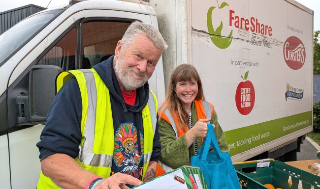 Photo of a man and a woman standing in front of a white van with FareShare South West logos, alongside others, on the side. The man is wearing a dark blue hoodie underneath a yellow high vis vest and holding a box. The woman is wearing a green fleece underneath an orange high vis vest and holding a blue shopping bag. Both are smiling at the camera and there is a green crate with oranges in just visible on the table in front of them