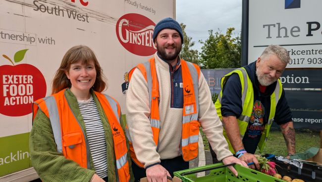 Photo of three people standing in front of a white van with FareShare South West logos, alongside others, on the side. The man on the right is wearing a dark blue hoodie underneath a yellow high vis vest and holding a box. The man in the middle is wearing a beige fleece underneath an orange high vis vest and a blue woolly hat. The woman on the left is wearing a green fleece underneath an orange high vis vest. All three are smiling at the camera and there is a green crate is just visible on the table in front of them. There is also the edge of The Park sign just visible to the right of the photo
