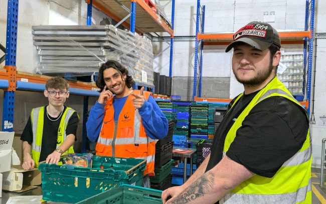 Three young men stand in a warehouse setting. The person on the far right is closest to the camera. He is wearing a black t-shirt under a yellow high vis vest and a black cap with a logo on. He is turned towards the camera with a smile and his hands are on a green crate. The middle man is wearing a blue jumper under an orange high vis vest. He is smiling at the camera and pointing. The person on the left is wearing a black t-shirt under a yellow high vis vest and resting his hands on a crate and a box as he smiles at the camera. There is shelving and stacks of crates in the background
