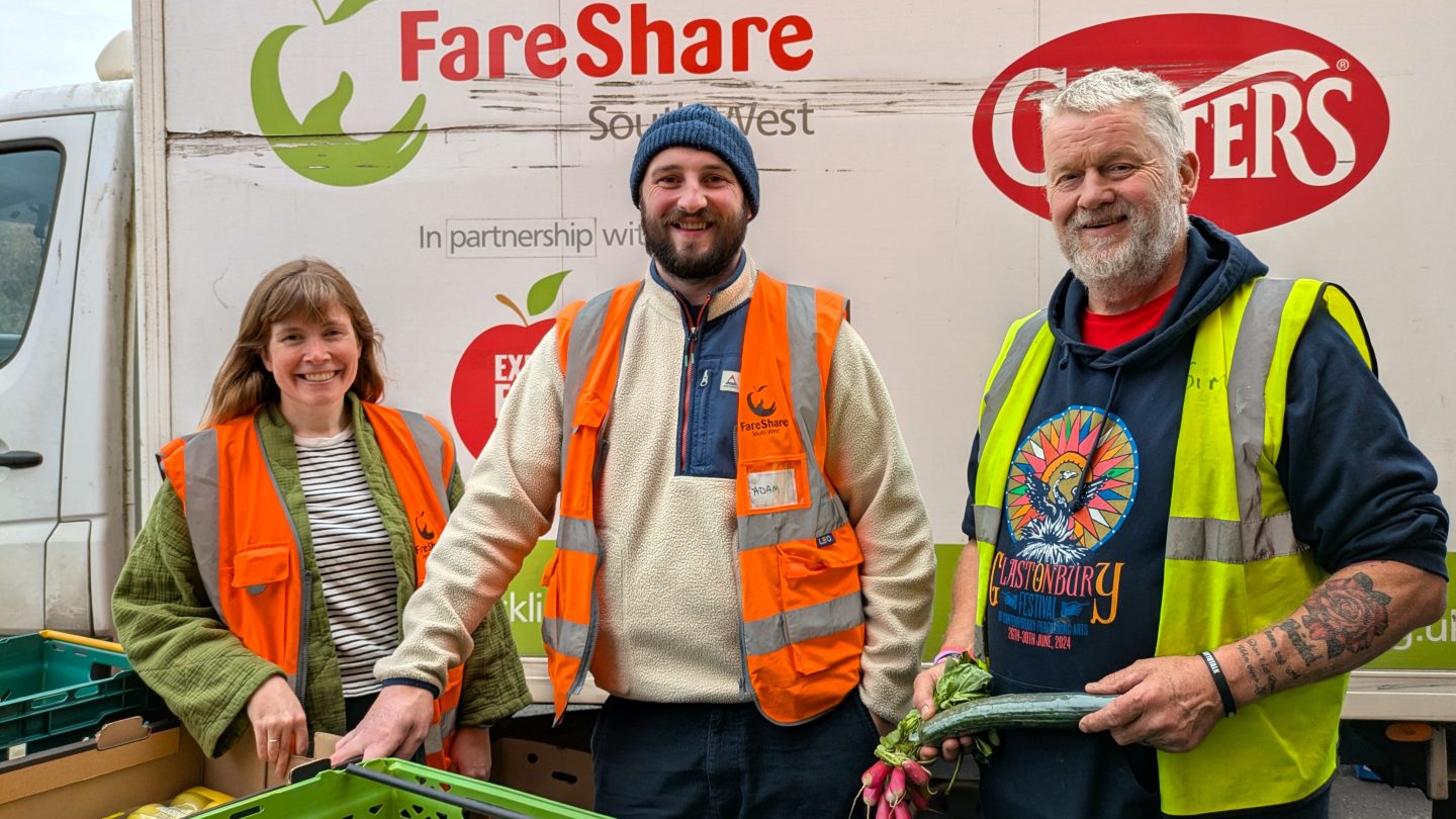 Photo of three people standing in front of a white van with FareShare South West logos, alongside others, on the side. The man on the right is wearing a dark blue hoodie underneath a yellow high vis vest and holding a box. The man in the middle is wearing a beige fleece underneath an orange high vis vest and a blue woolly hat. The woman on the left is wearing a green fleece underneath an orange high vis vest. All three are smiling at the camera and there is a green crate is just visible on the table in front of them