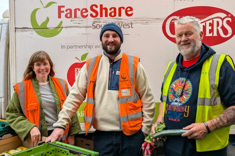 Photo of three people standing in front of a white van with FareShare South West logos, alongside others, on the side. The man on the right is wearing a dark blue hoodie underneath a yellow high vis vest and holding a box. The man in the middle is wearing a beige fleece underneath an orange high vis vest and a blue woolly hat. The woman on the left is wearing a green fleece underneath an orange high vis vest. All three are smiling at the camera and there is a green crate is just visible on the table in front of them