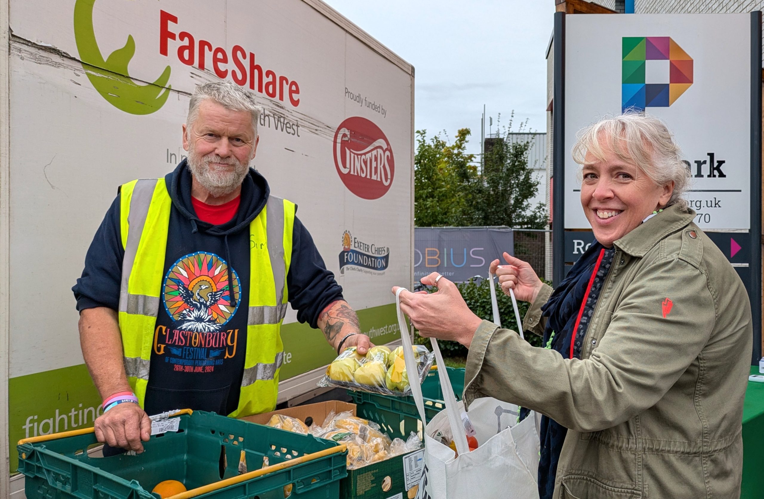 Photo of a man and a woman standing in front of a white van with FareShare South West logos, alongside others, on the side. The man is wearing a dark blue hoodie underneath a yellow high vis vest and smiling at the camera. The woman is on the other side of a table and wearing a green coat. She is holding a shopping bag as she smiles at the camera. There are rows of green crates full of fruit and veg between them. In the background to the right of the photo is the edge of a building and a large sign on which   the end of the word 'The Park' is just visible