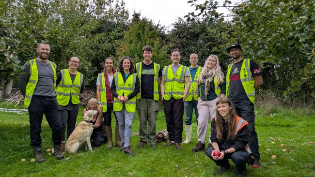 A group of 11 people are gathered in an orchard. All but two are wearing yellow high vis vests. The person at the far right is crouching down wearing an orange high vis vest and holding an apple. The person third from the left is crouching behind a golden retriever dog and wearing a black vest. All are smiling at the camera. There is grass in the foreground and trees beyond