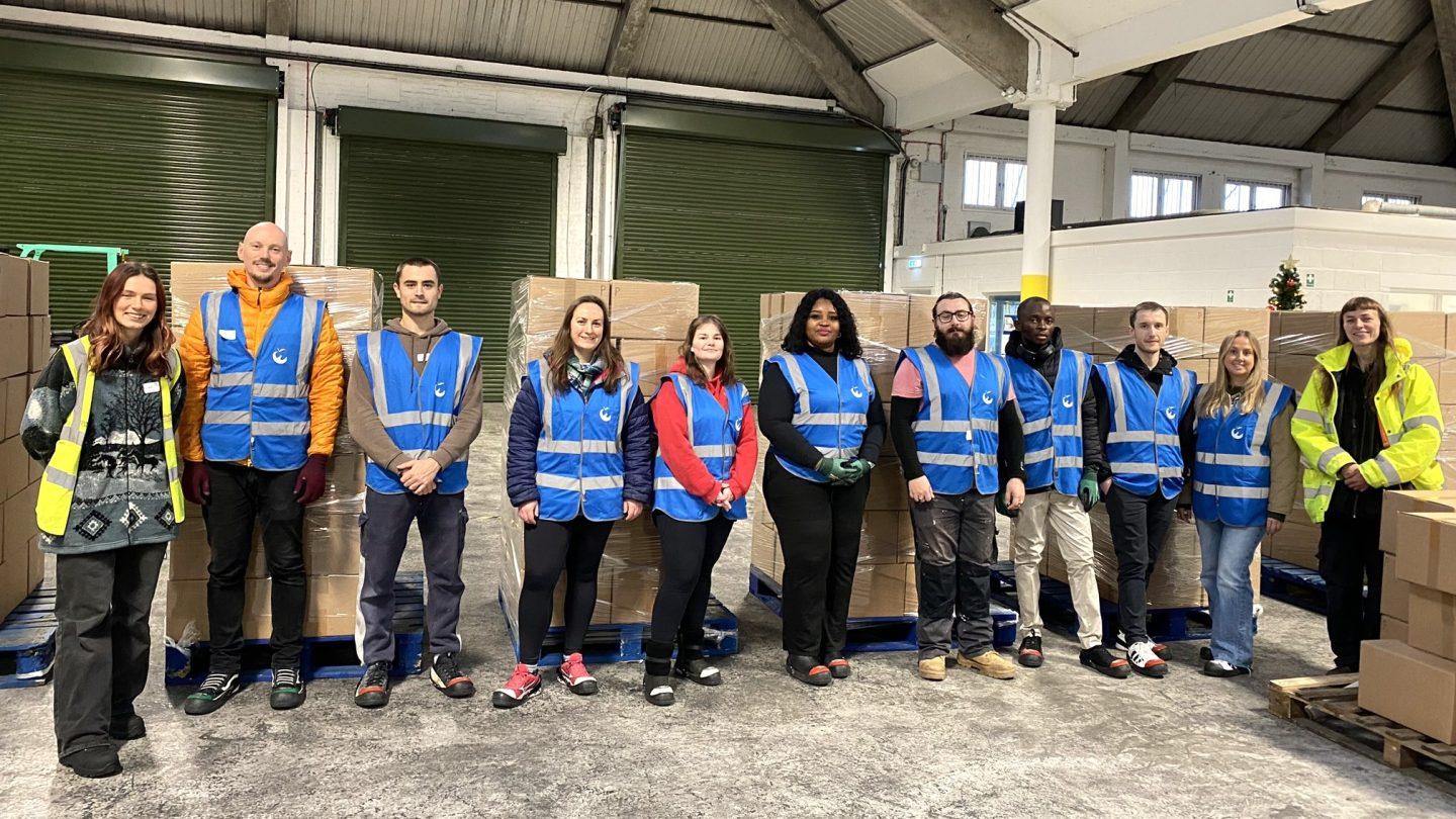 Group shot of 11 people standing in a row. The person on the far left is wearing a yellow high vis vest and the person on the far right is wearing a yellow high vis jacket. Everyone else is wearing a blue high vis jacket over warm winter clothes. They are standing in a warehouse and are surrounded by stacks of cardboard boxes