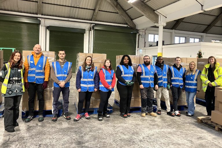 Group shot of 11 people standing in a row. The person on the far left is wearing a yellow high vis vest and the person on the far right is wearing a yellow high vis jacket. Everyone else is wearing a blue high vis jacket over warm winter clothes. They are standing in a warehouse and are surrounded by stacks of cardboard boxes