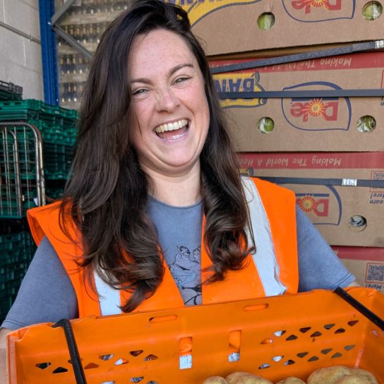 Person with long dark hair laughs as she holds an orange crate full of potatoes. She is looking towards the camera and is wearing a blue top underneath an orange high vis vest. There are cardboard boxes stacked behind her in a warehouse setting