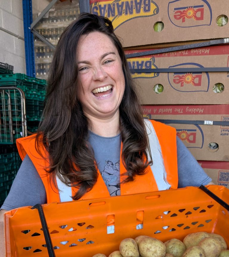 Person with long dark hair laughs as she holds an orange crate full of potatoes. She is looking towards the camera and is wearing a blue top underneath an orange high vis vest. There are cardboard boxes stacked behind her in a warehouse setting