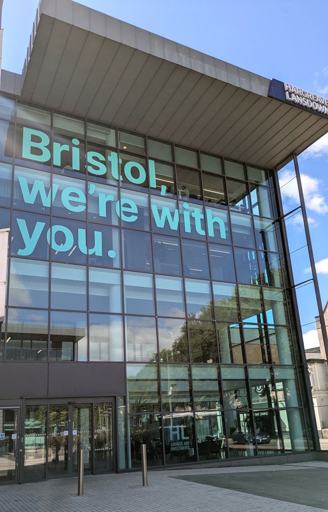 Close up of a large glass building. Large letters on the front say: Bristol, we're with you. The Hargreaves Lansdown logo is at the top right of the building and there is blue sky in the background