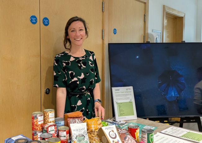 A woman with brown hair wearing a green dress with pink and black print is smiling at the camera. She stands behind a desk with items of food, including cans and packets, and a couple of sheets of paper with the FareShare South West logo on and 'winter food collection' written on. There is a large, blank TV screen to one side of her and brown wooden cupboards behind