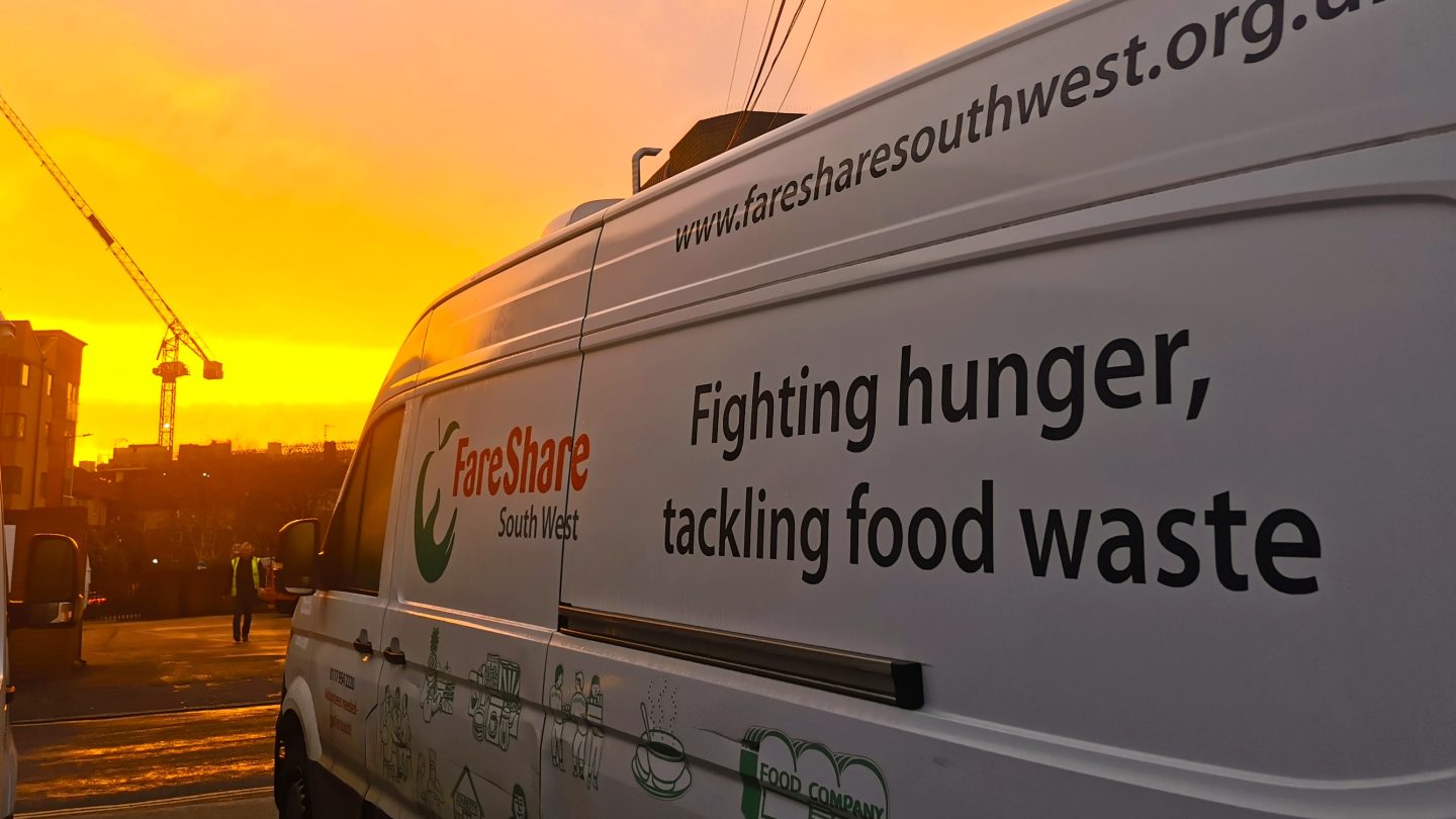 Close up of a FareShare South West-branded white van with a yellow and orange sunrise in the background and a crane silhouetted in the distance